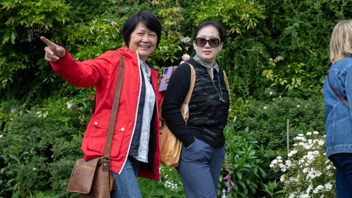 Two smiling visitors walk in front of lush green vegetation, white and purple flowers in Crook Hall Gardens